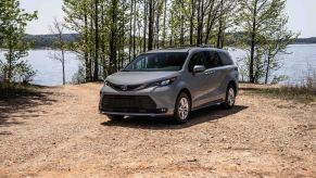 A silver Toyota Sienna in a dirt area with trees and a body of water in the background.