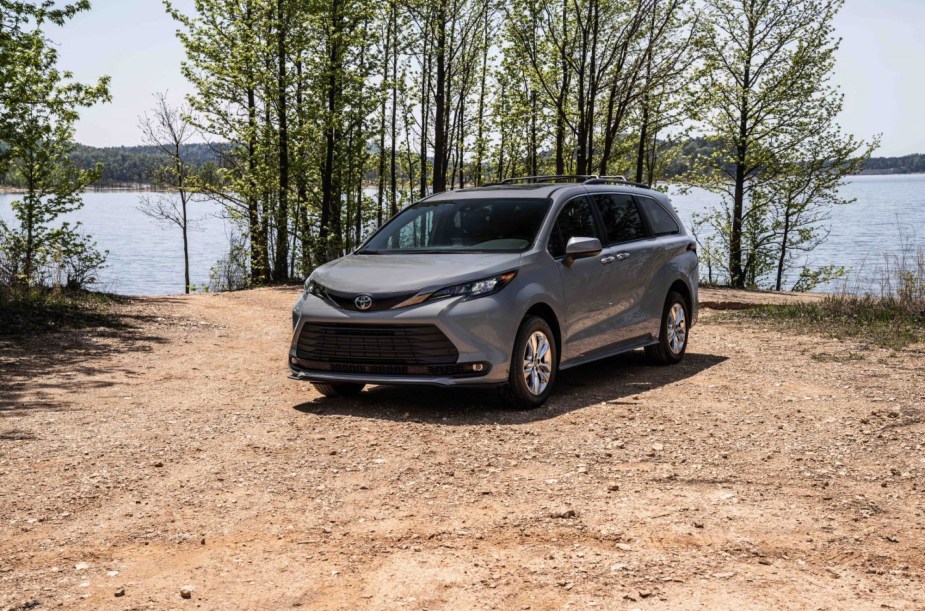 A silver Toyota Sienna in a dirt area with trees and a body of water in the background. 