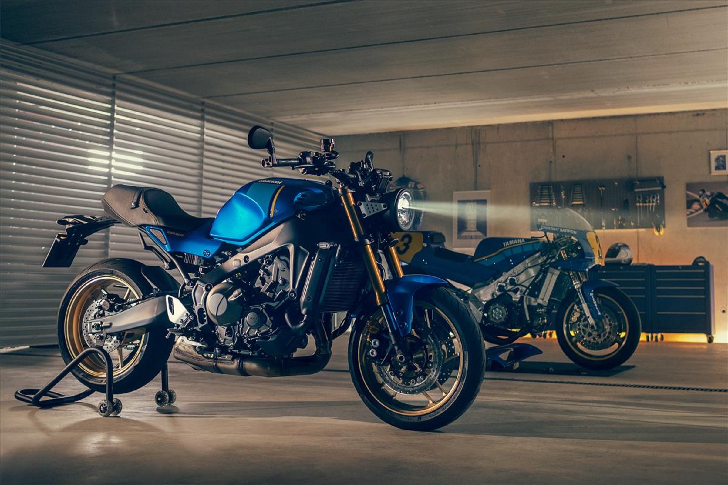 A blue-and-black 2022 Yamaha XSR900 in a garage in front of a blue 1980s Yamaha GP race bike