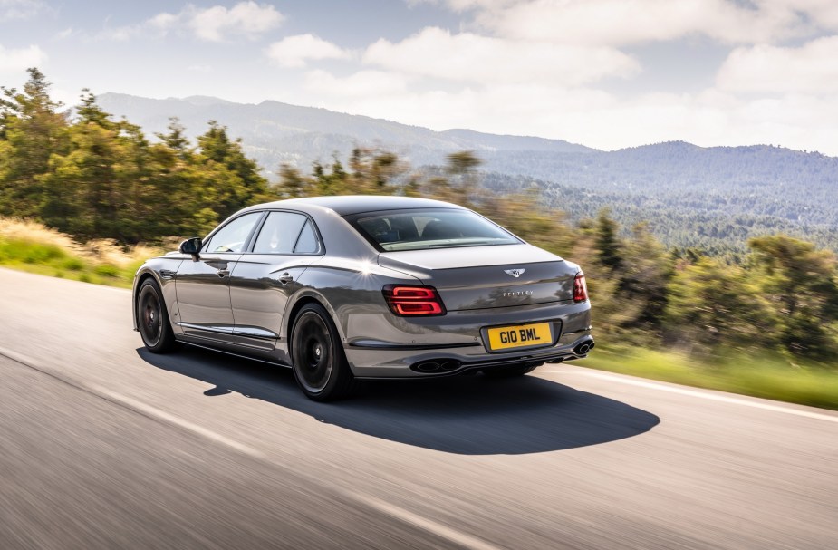 The rear 3/4 view of a gray 2023 Bentley Flying Spur S driving down a European mountain forest road