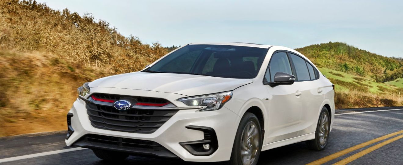 The refreshed white 2023 Subaru Legacy midsize sedan model driving down a country highway near grassy hills