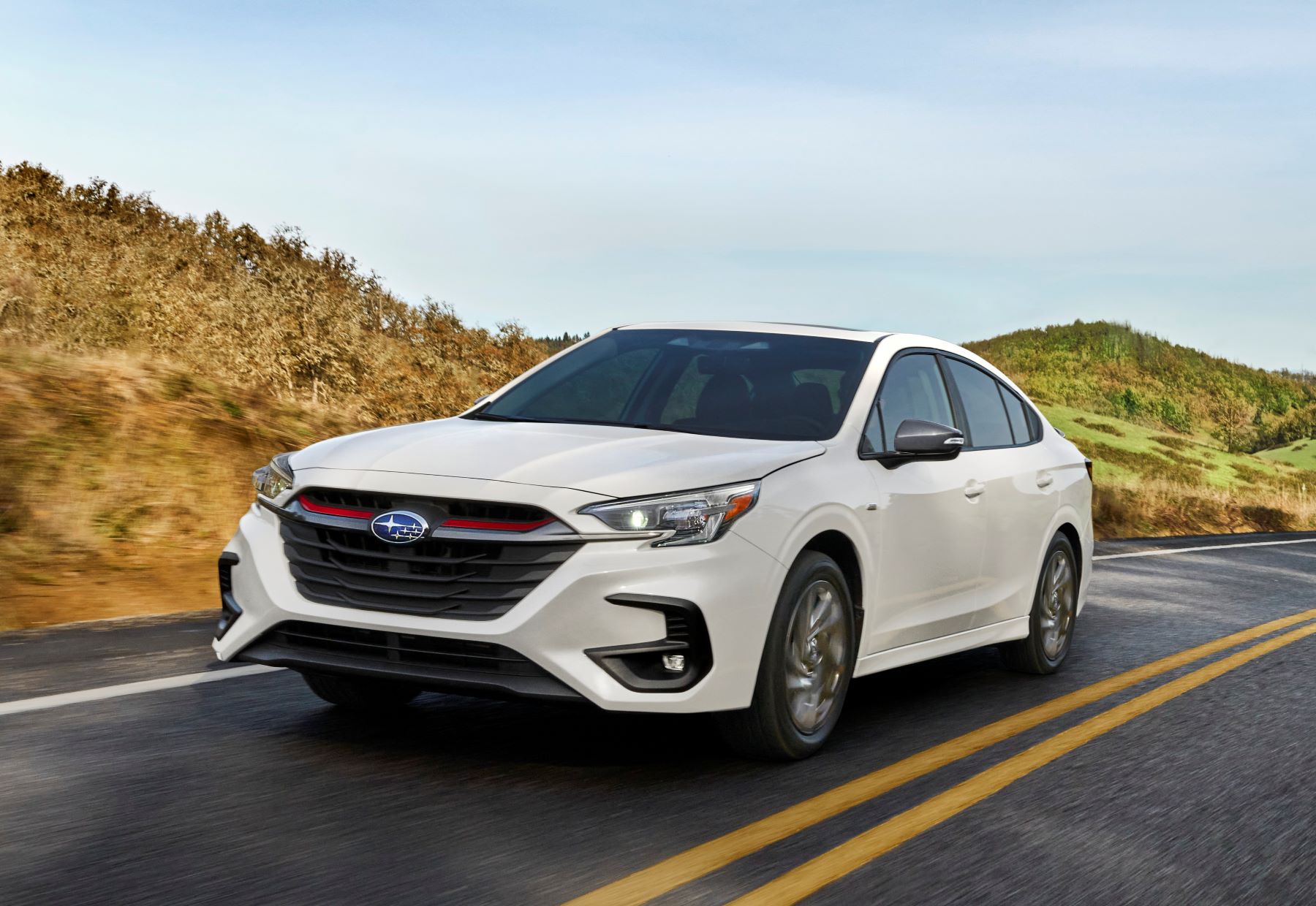 The refreshed white 2023 Subaru Legacy midsize sedan model driving down a country highway near grassy hills