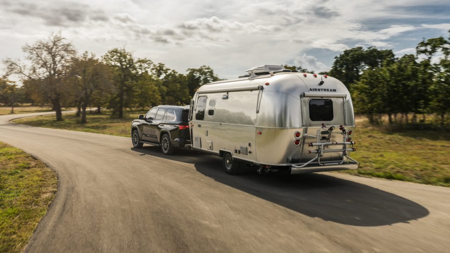 2023 Toyota Sequoia SUV towing an airstream camper trailer down a curvy, tree-lined road.