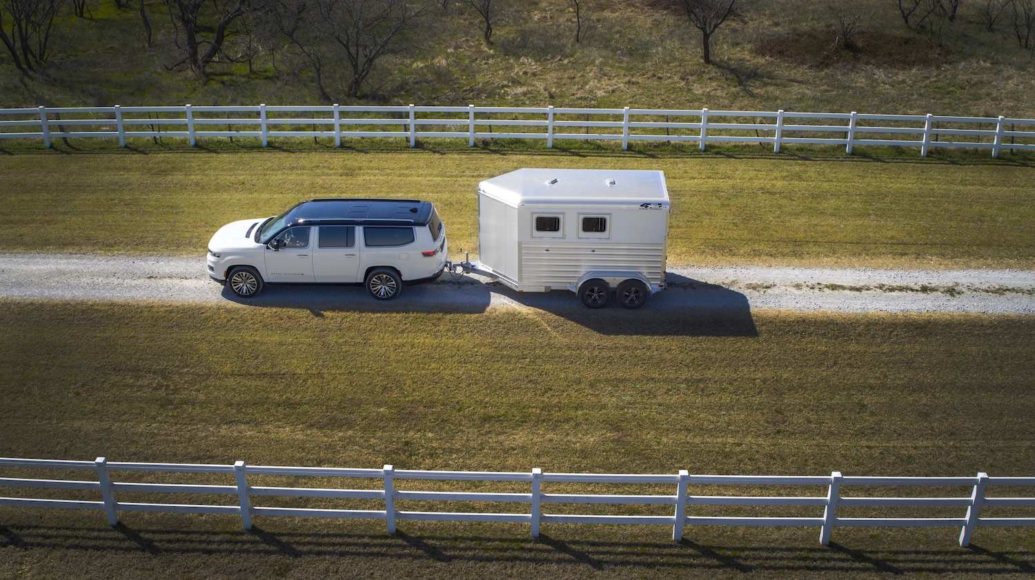 Overhead view of a white Grand Wagoneer SUV towing a horse trailer down a driveway, white fences visible on both sides.
