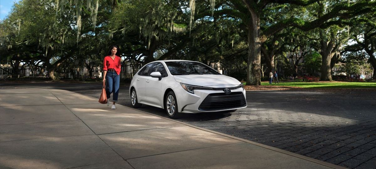 Woman in red holding a coffee walking toward a new white 2023 Toyota Corolla sedan parked on a street