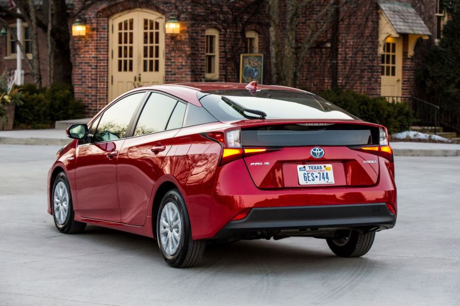 Rear angle view of a fourth-generation Toyota Prius in red, parked in front of a fancy brick house.