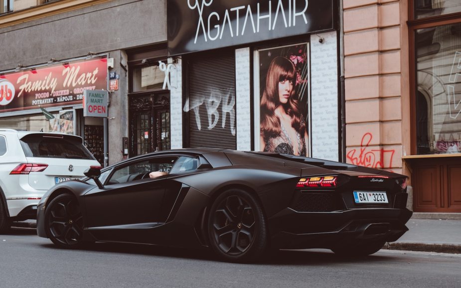 A black Lamborghini parked on a city street, a salon and other businesses visible in the background.