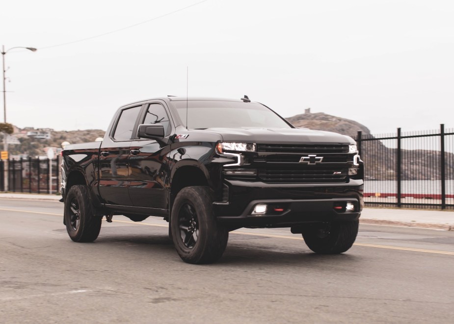 Black Chevrolet Silverado pickup truck driving along a harbor, the ocean and mountains visible in the background.