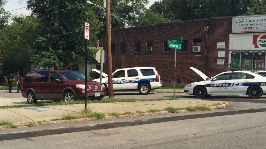 Two police cruisers parked on a side street with their hoods up, a brick walled convenience store visible behind them.
