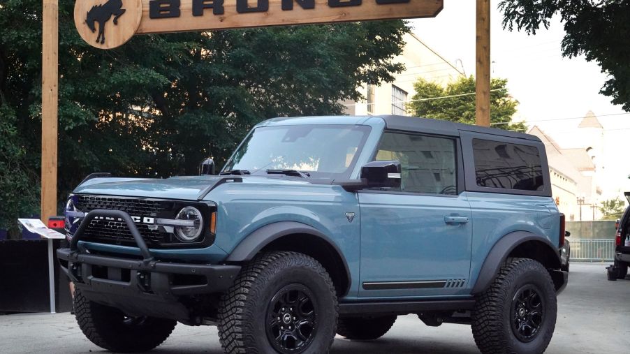 A light blue Ford Bronco parked outdoors in front of a wooden Bronco sign.