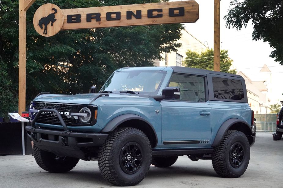 A light blue Ford Bronco parked outside in front of a wooden Bronco sign. 