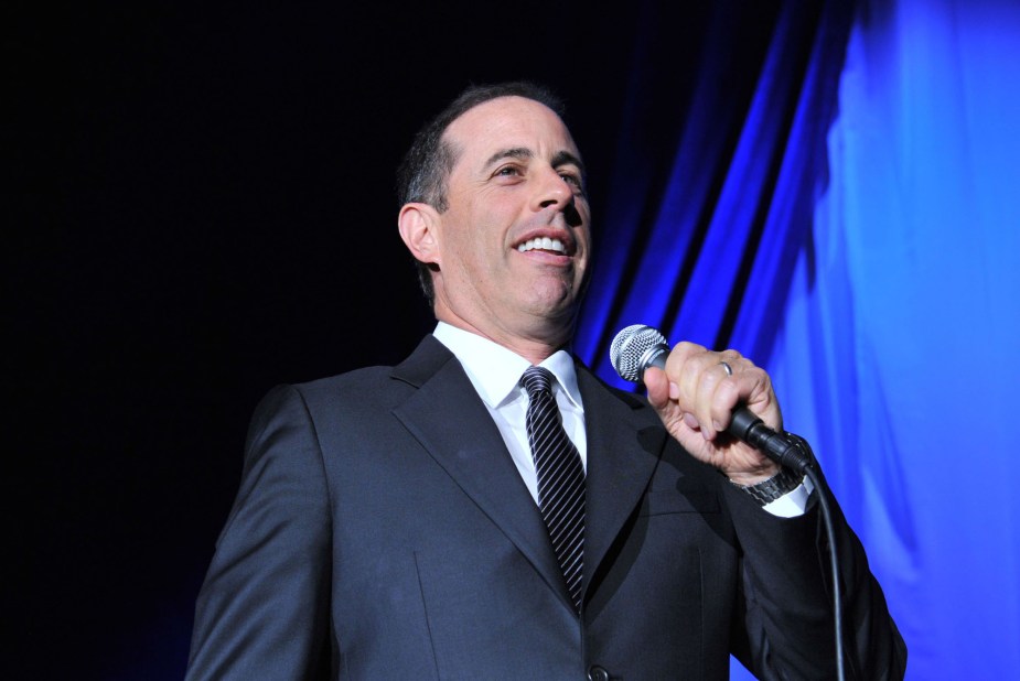 Jerry Seinfeld on a stage with blue background.