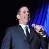Jerry Seinfeld on a stage with blue background.