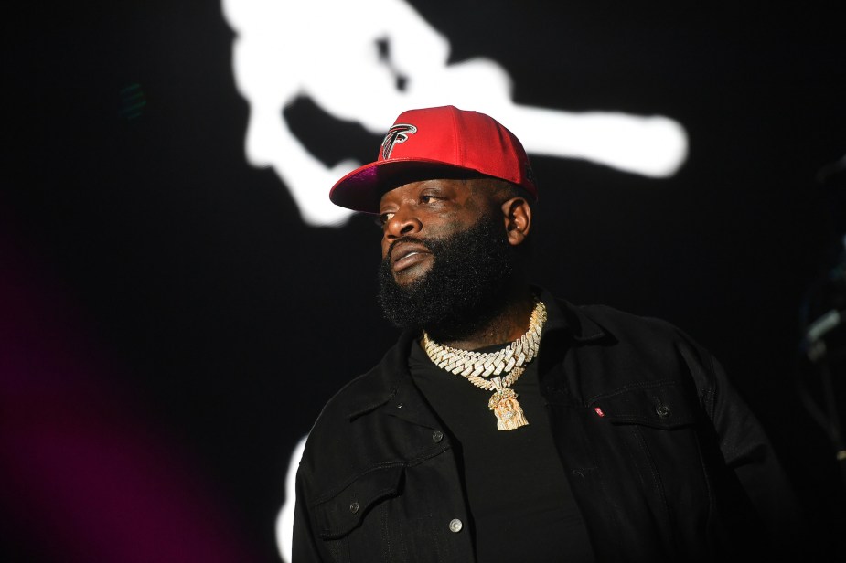 Rick Ross dressed in black in front of black background with a red hat.
