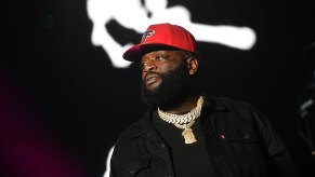Rick Ross dressed in black in front of black background with a red hat.