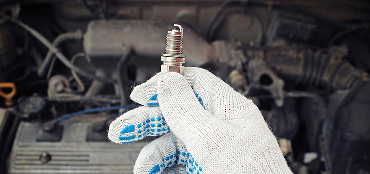A gloved mechanic's hand holding a new spark plug over an older engine.