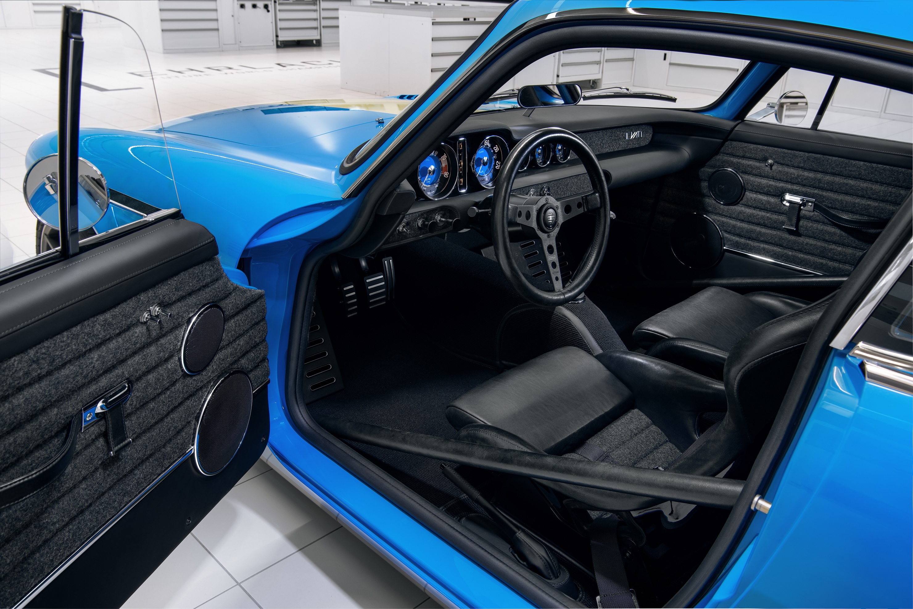The black Recaro seats, roll cage, and dashboard of a blue Volvo P1800 Cyan in a white garage