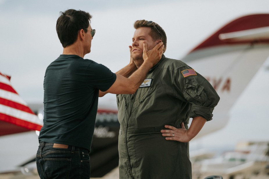Tom Cruise and James Corden standing out side with Cruise's hands on Corden's face in front of a jet. 