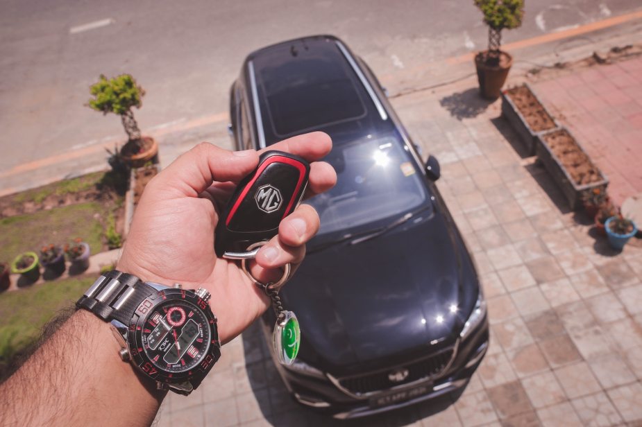 Closeup of a man's hand holding a car key fob, the crossover SUV available in their driveway, in the background.