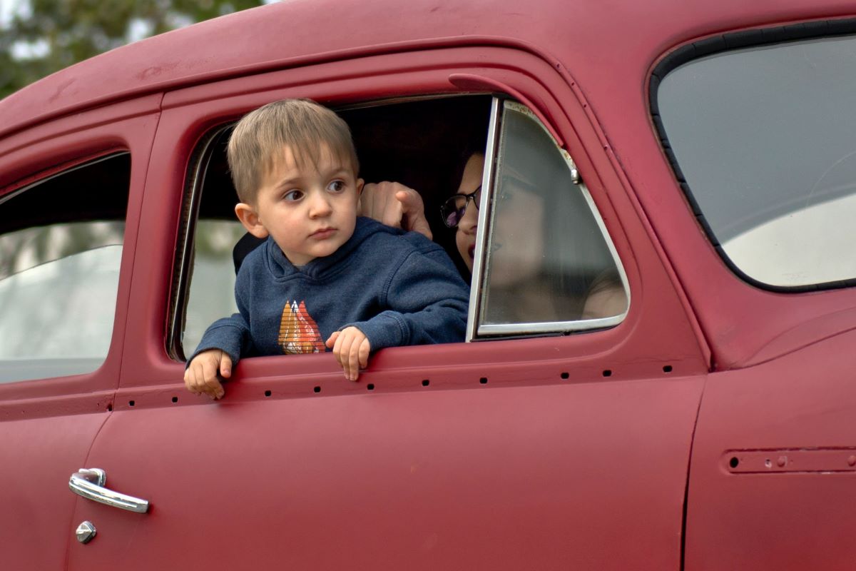 A child in a blue sweatshirt leans out the open window of an older model year red car