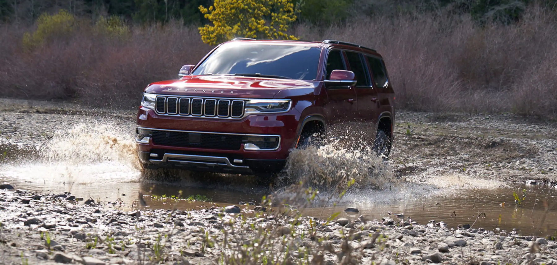 A dark red 2022 Jeep Wagoneer full-size luxury SUV driving through a creek in the wilderness