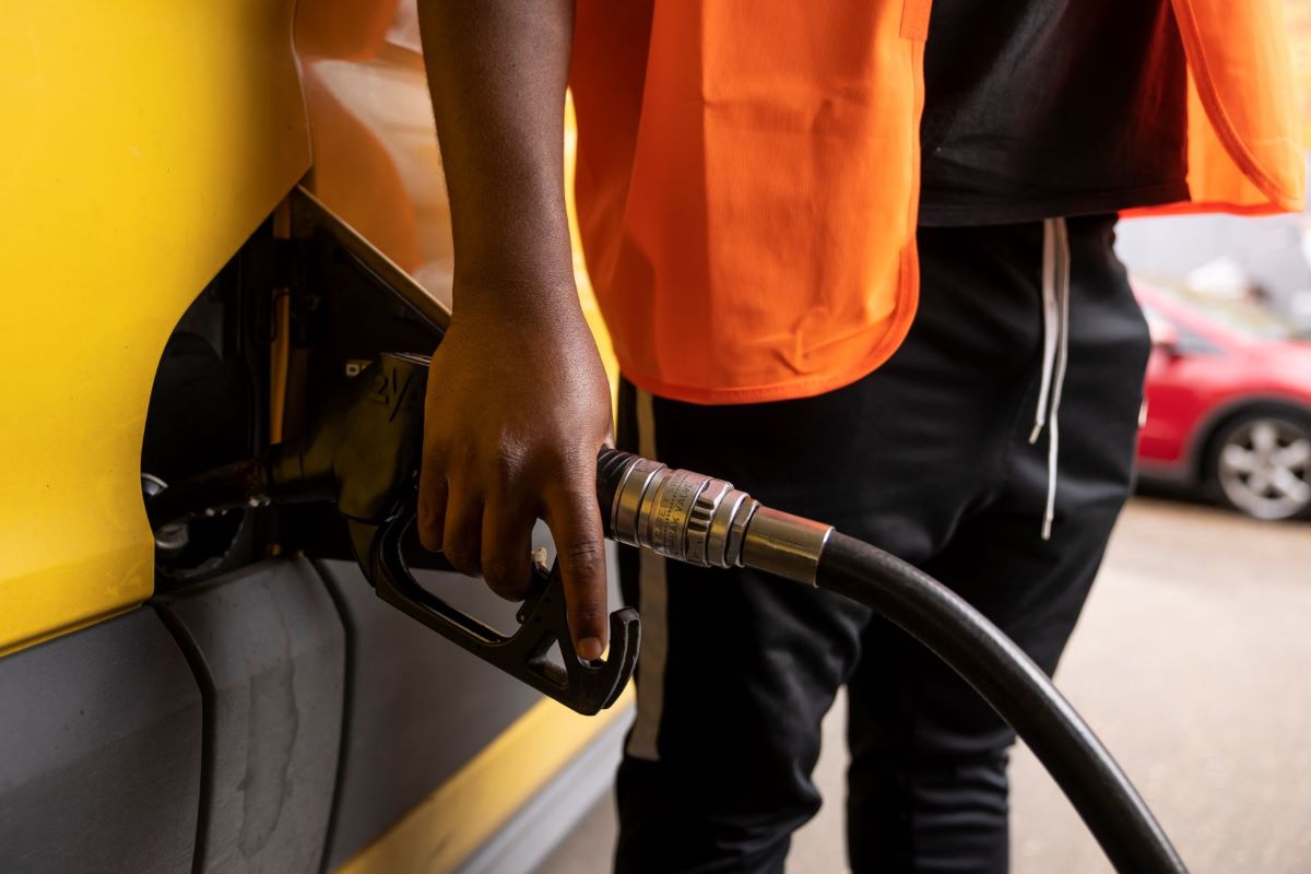 A Black man in stylish clothing holds onto a fuel pump as he adds gas to his gas tank