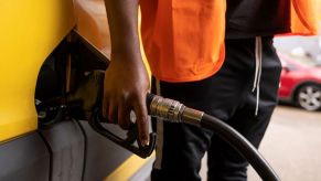 A Black man in stylish clothing holds onto a fuel pump as he adds gas to his gas tank