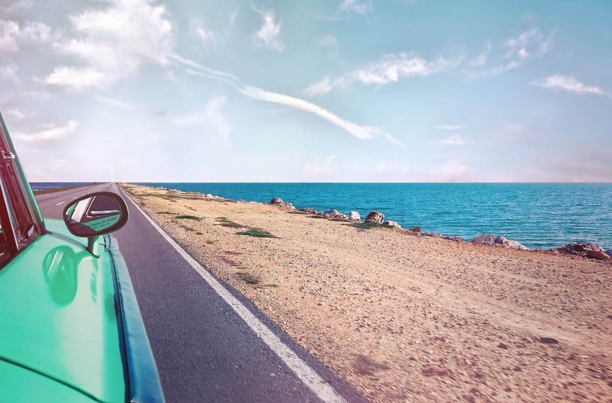 A teal classic car driving along a road next to the ocean on a hot summer day