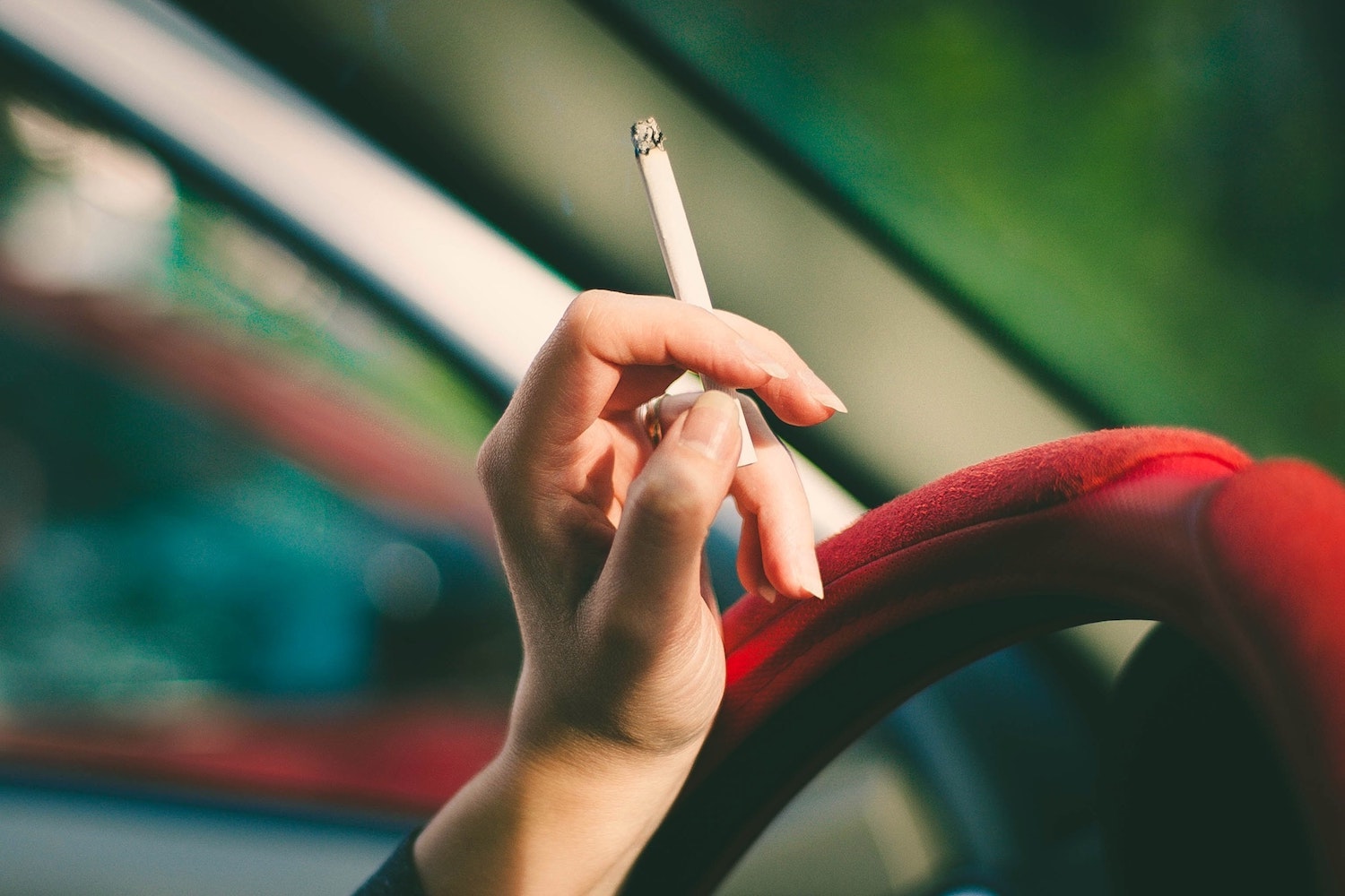 Closeup of a woman's hand holding a cigarette against the steering wheel of her car.