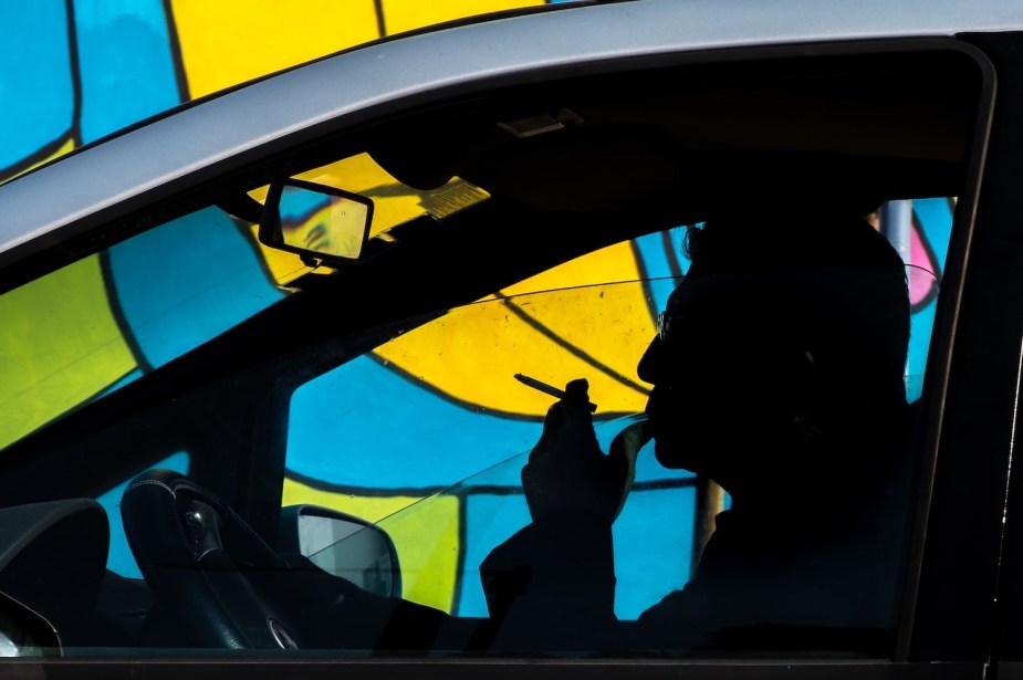 Silhouette of a man smoking a cigarette in the driver's seat of his parked car, a colorful mural visible in the background.