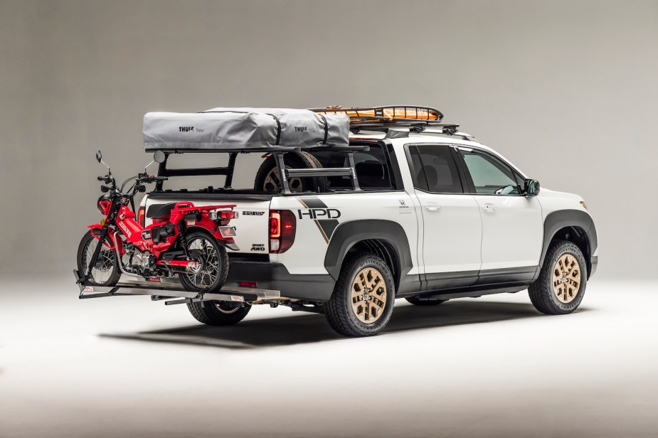 White Honda Ridgeline pickup truck hauling a rooftop tent, roof rack, and a motorcycle on its trailer hitch bike carrier.