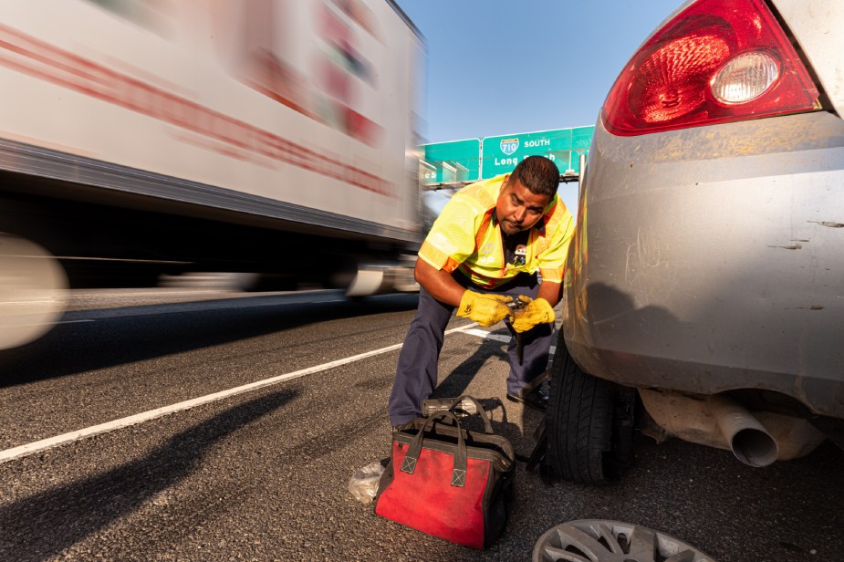 A Freeway Service Patrol worker in a high-vis vest tries to change a flat tire on the side of the road as a semi-truck passes