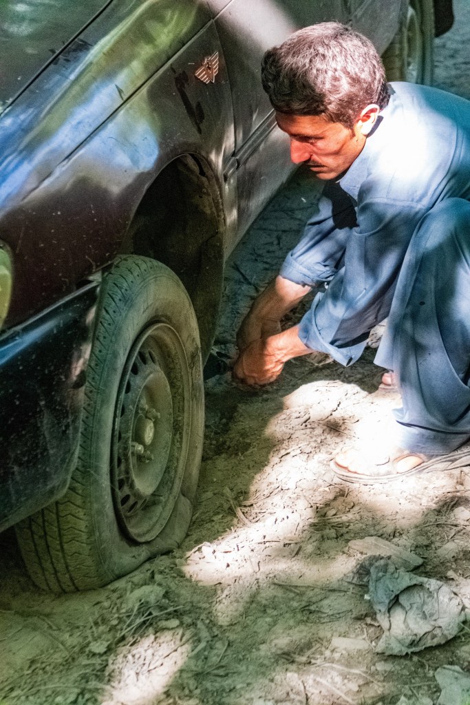 A blue mechanic tries to change a car's flat tire in the dirt