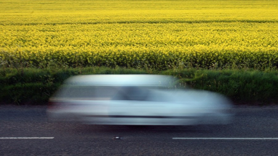 A white car whizzing down a country road at high speed appears as a blur in front of yellow flowers.
