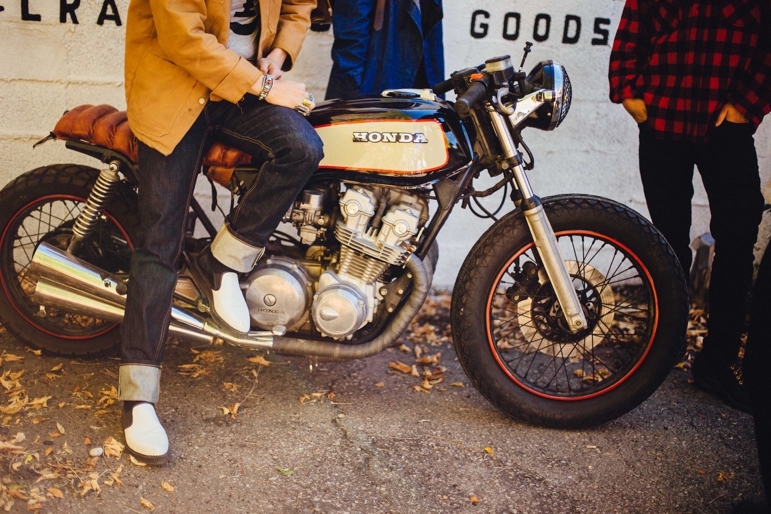 Customized cafe racer motorcycle parked by a brick wall, its rider sitting atop its seat, two other people's legs visible nearby.