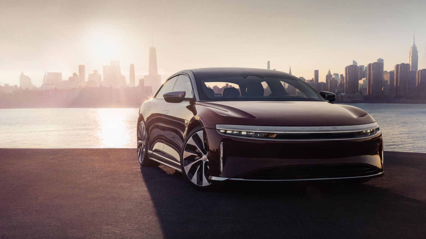 A 2022 Lucid Air luxury electric sedan model parked by the sea as the sun rises of a city skyline of skyscrapers