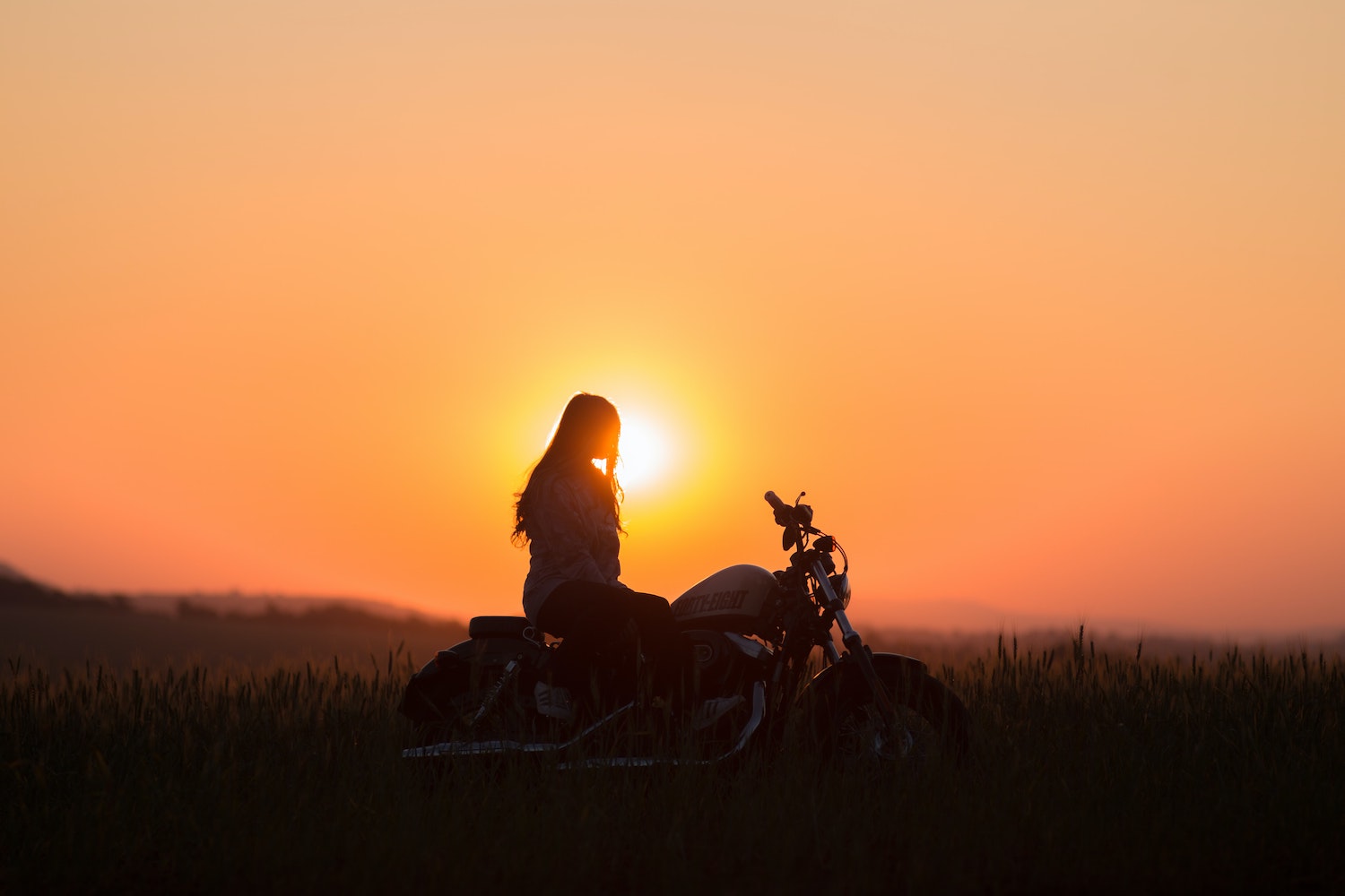 Woman sitting atop a Harley Davidson motorcycle while watching the sun set, a grassy field visible in the background.
