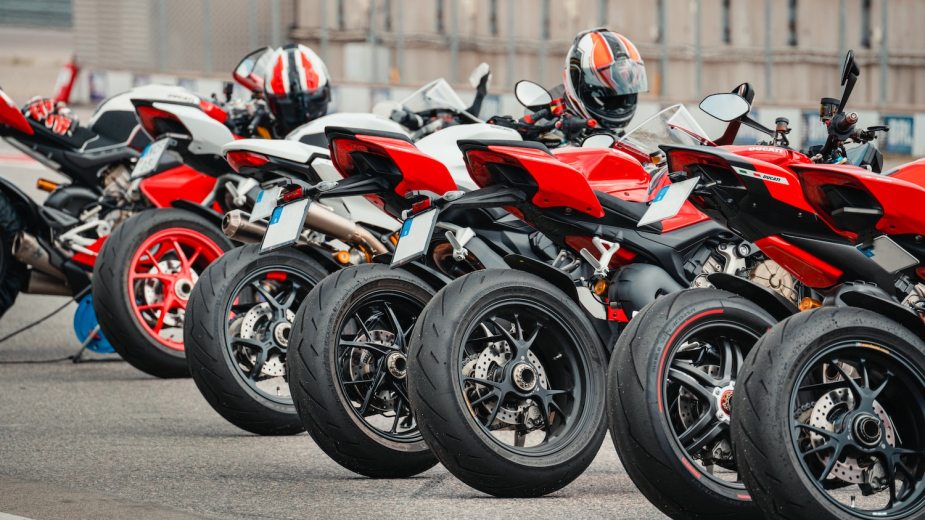 A row of red and white sport bike motorcycles parked at a race track, a fence visible in the background.
