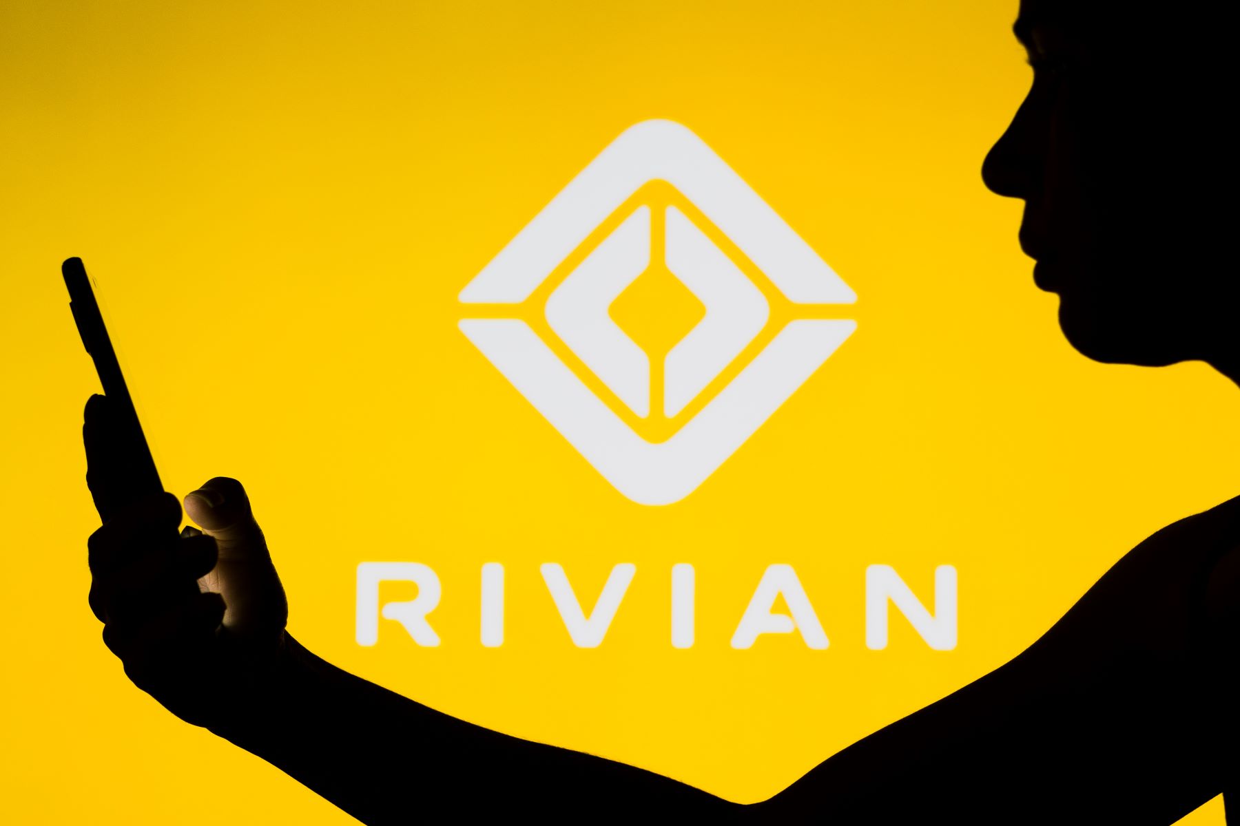 The Rivian EV company logo with a silhouette of someone on a smartphone in the foreground