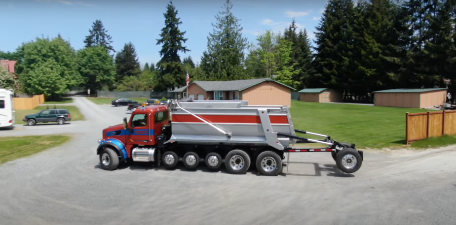 Red dump truck deploying a drop axle and two wheels, a suburban street visible in the background.
