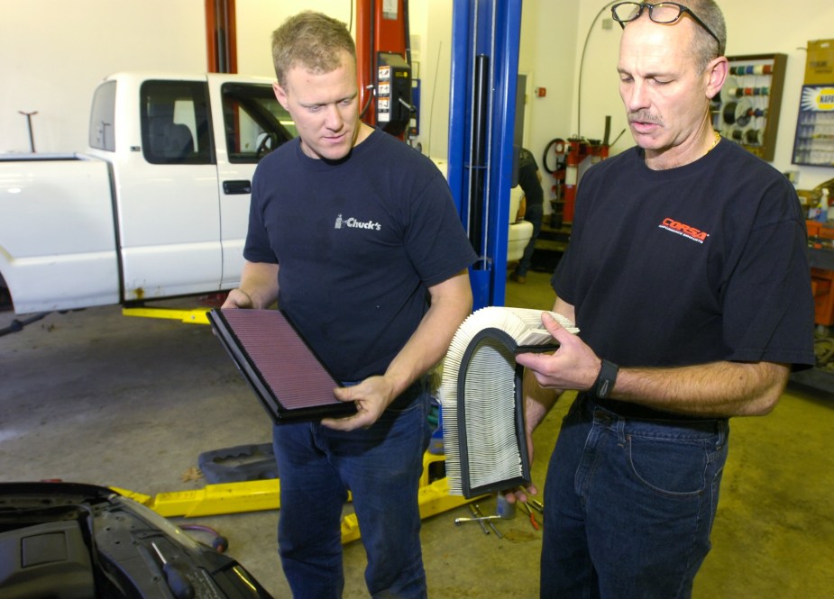Two car mechanics in a garage compare a stock white (right) and high-performance red engine air filter