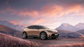 A bronze 2023 Genesis GV60 electric SUV model parked in a mountain field bathed in light from a purple cloudy sky