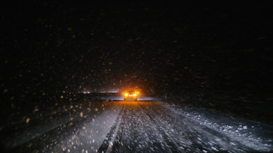 A car with its hazard lights on is driving through heavy snow flurries.