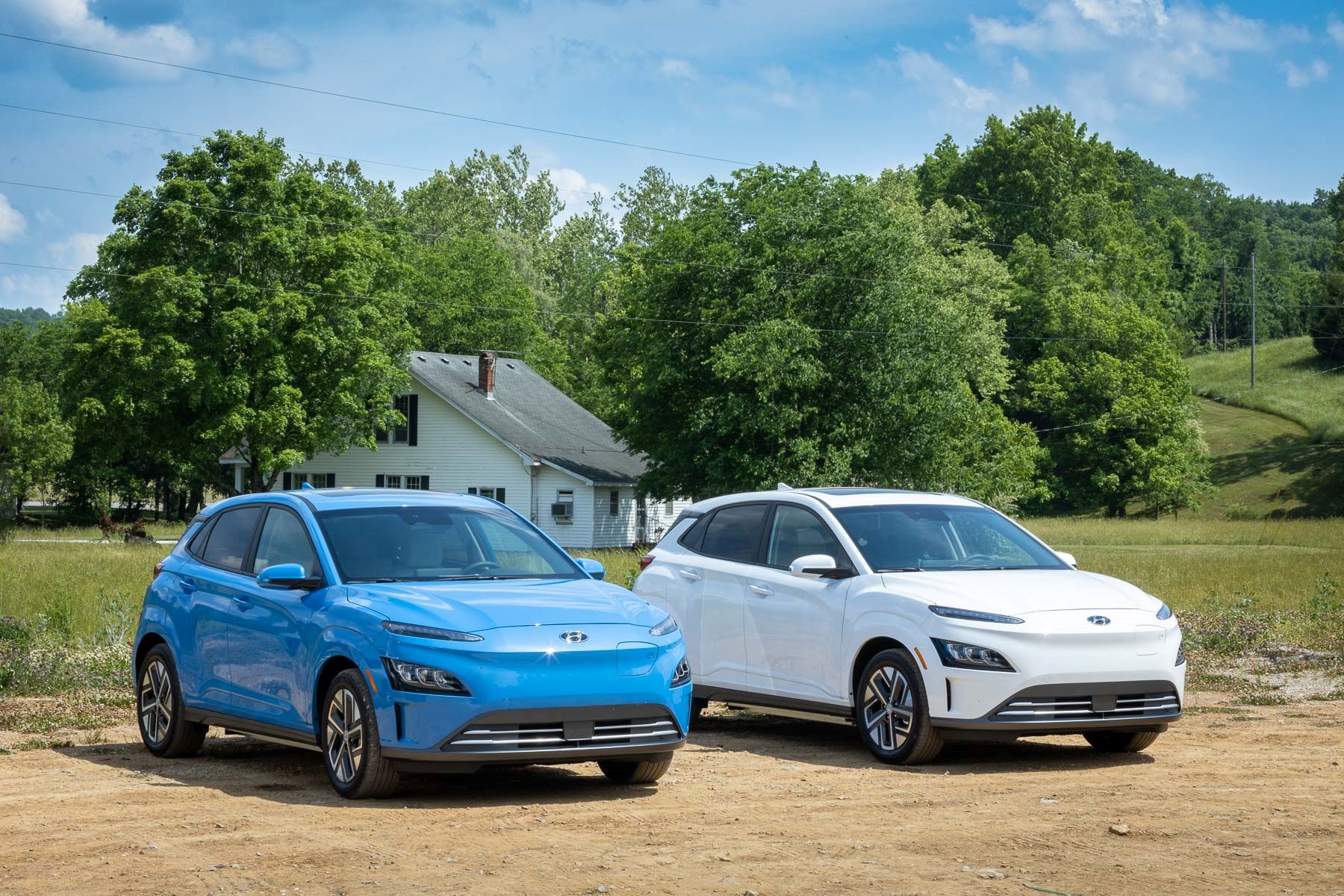Light blue and white 2022 Hyundai Kona Electric SUV models parked on a dirt path in front of a house surrounded by trees