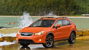 An orange 2013 Subaru XV Crosstrek compact SUV parked on a wet pier near splashing washes from a forest lake
