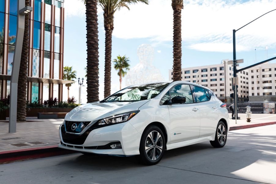 A white 2019 Nissan Leaf Plus EV hatchback model parked near a cluster of palm trees on a city street
