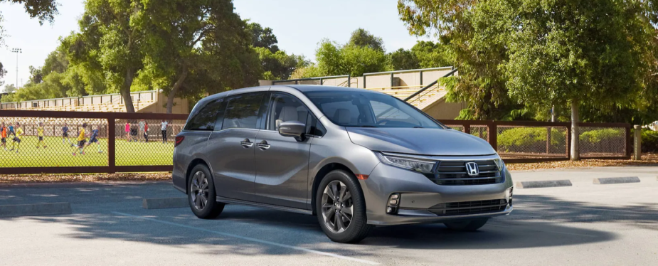 A silver gray 2023 Honda Odyssey minivan parked near a sports field where children are playing soccer