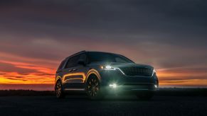 A 2023 Kia Carnival minivan model parked in a field at sunset with front headlights turned on
