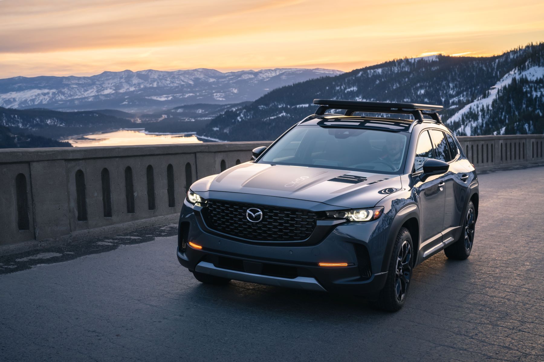A 2023 Mazda CX-50 parked on a bridge over a river near snowy mountains as the sun sets
