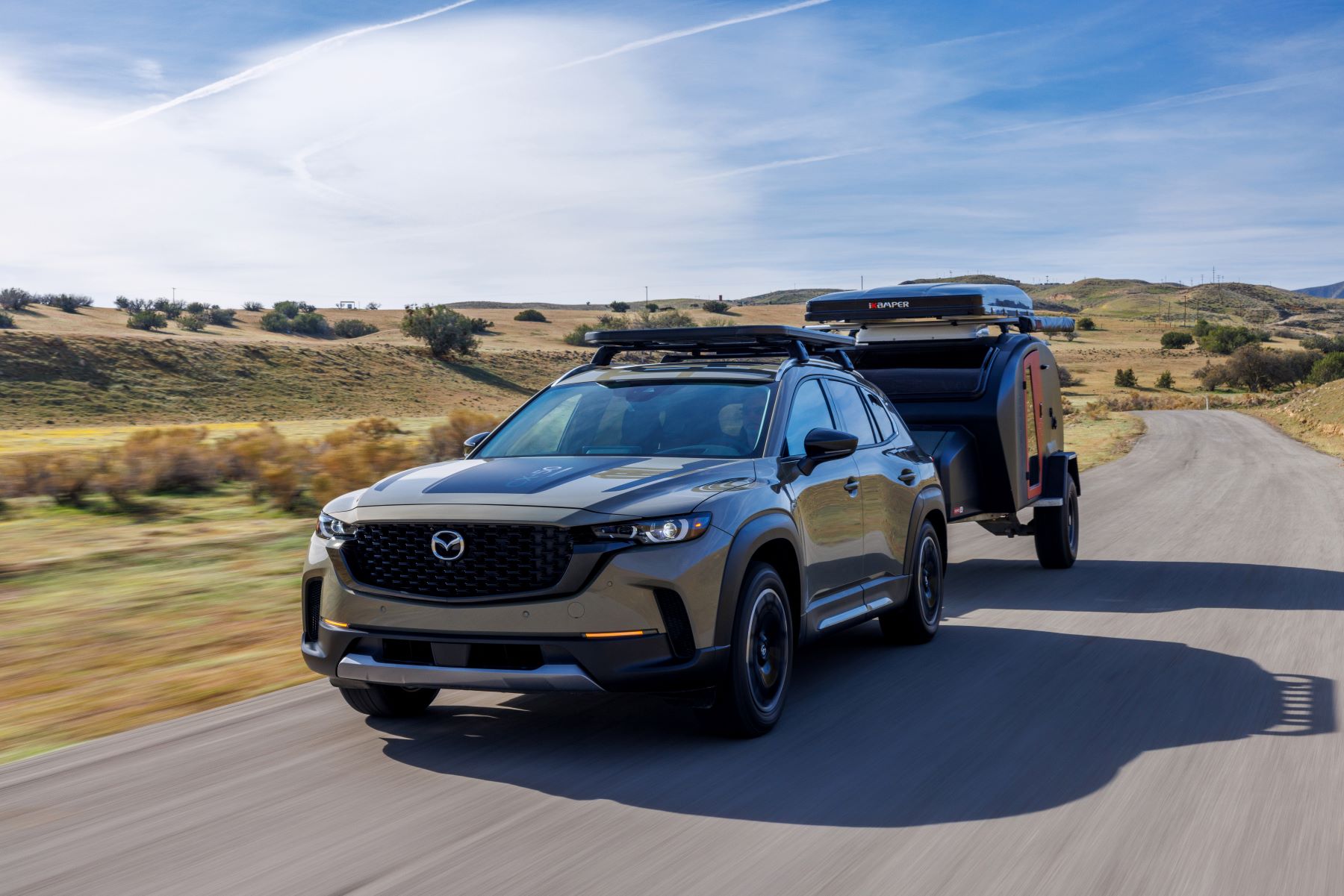 A 2023 Mazda CX-50 compact crossover SUV towing a travel trailer on a country highway
